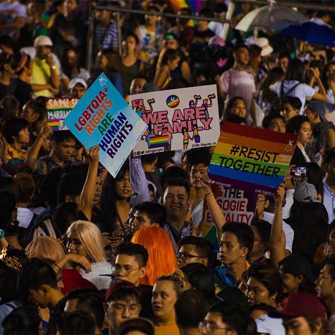 Image description: A crowd of people from the 2019 Pride March and Festival. In the center, there is a smiling person raising a placard that says 'WE ARE FAMILY' in pink block text. Various queer couples waving rainbow flags around the text. To their right, a person is raising a placard that says '#RESIST TOGETHER' with a rainbow background. On the left side, another person is holding up a placard that says 'LGBTQIA+ rights are human rights' in block rainbow text and a sky background.